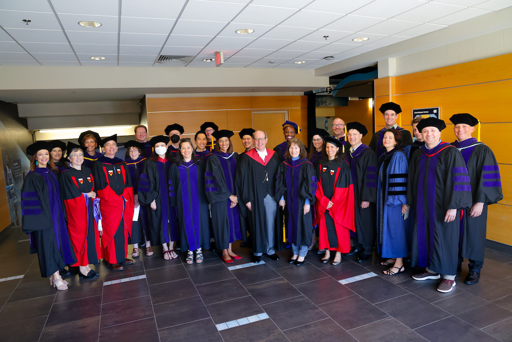 Faculty dressed in regalia pose together with Dean Martin Brinkley and commencement speaker Sydney Batch in the lobby of Carmichael arena.